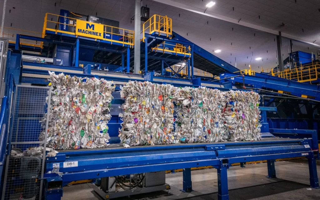 - RobotsInc.com Industrial recycling facility with blue machinery and large bales of compressed paper in the foreground