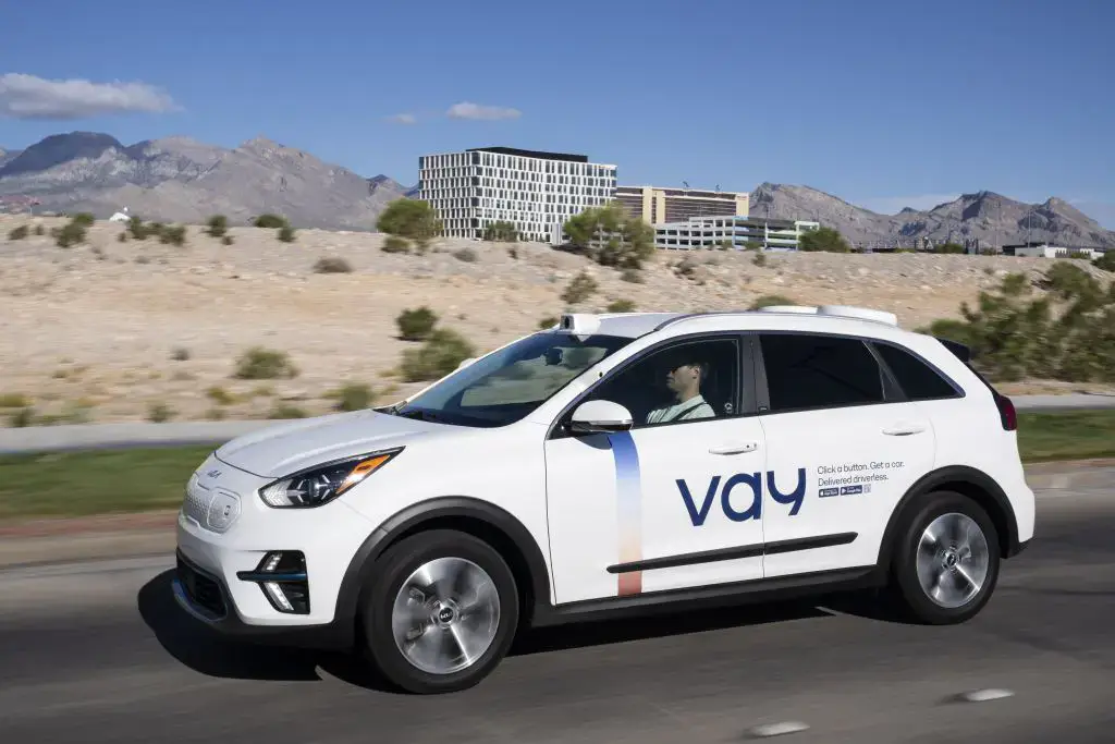 - RobotsInc.com White SUV with a blue 'vay' logo driving on a desert highway with mountains in the background, under a clear blue sky.