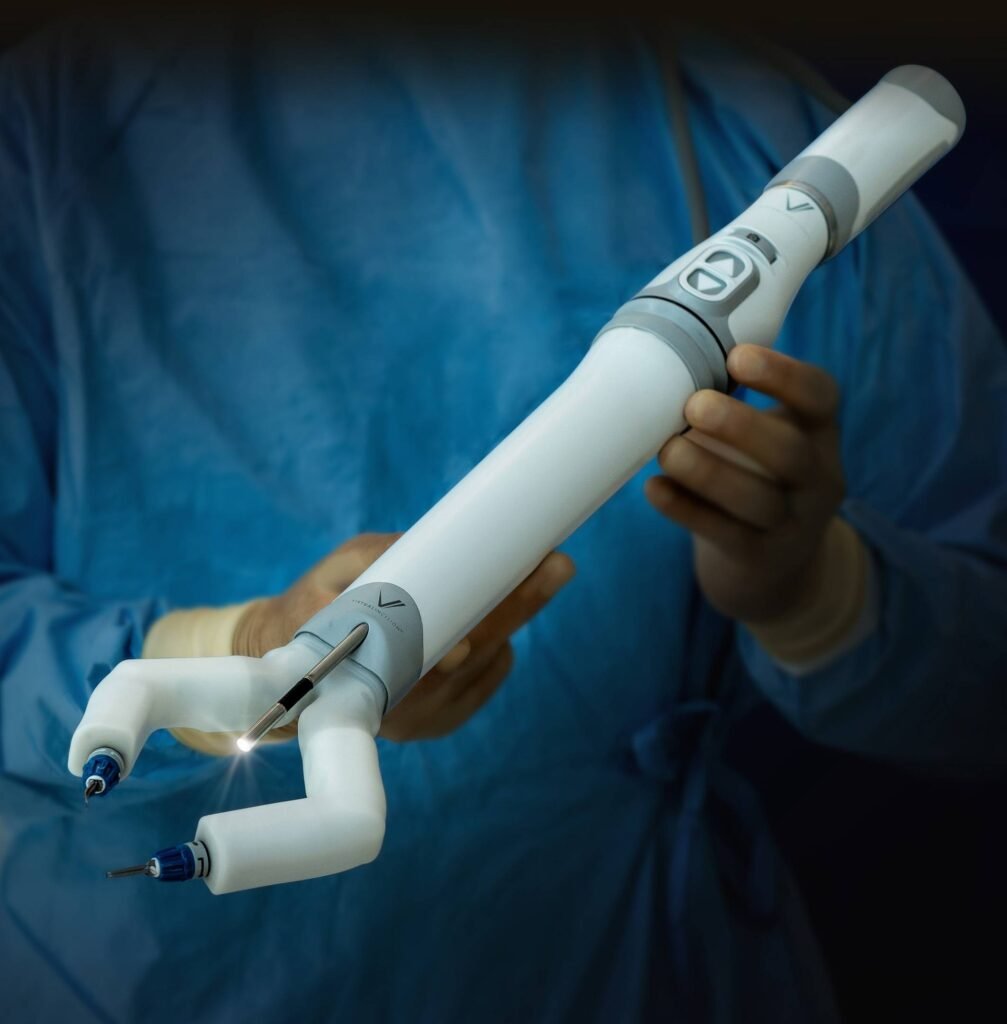- RobotsInc.com Close-up of a robotic surgical instrument held by gloved hands in a blue-surgical setting, with a lit tip on the instrument's end-effector.