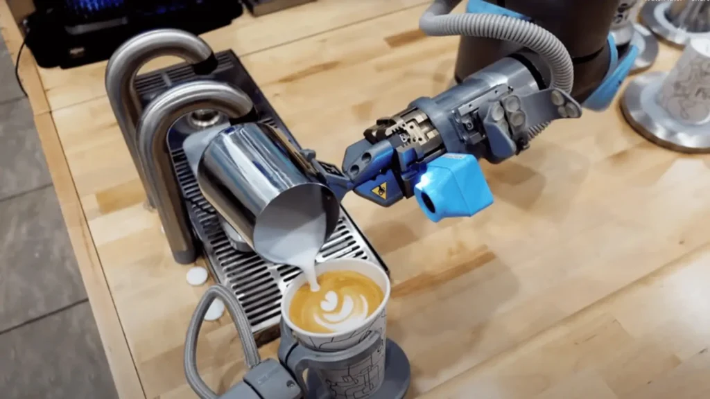 - RobotsInc.com Robot barista pours latte art into a cup of coffee at a counter while a robotic arm operates nearby.
