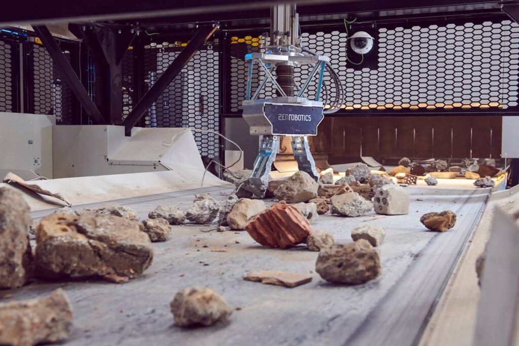 - RobotsInc.com Industrial robot with a blue frame and ‘ZEN ROBOTICS’ logo stands over a rock-filled conveyor in a lab-like testing area.